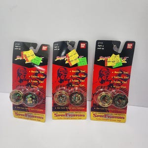 May include: Three Spin Fighters turbo-charged spinner tops on their original packaging. The packaging is red and yellow with the text "Spin Fighters" and "Die-Cast Metal Spin Fighters". Each package includes two metal spinners.