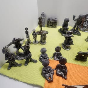 Może przedstawiać: Kolekcja figurek cynowych, w tym wieża zegarowa, dom, dziewczynka trzymająca lalkę, chłopiec trzymający psa i kilka innych figurek. Figurki są ułożone na zielonej i pomarańczowej powierzchni.