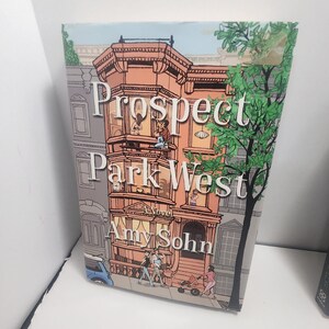 May include: A book titled "Prospect Park West" by Amy Sohn. The cover features an illustration of a brownstone building with people on the balconies and street. A blue car and a stroller are also visible.