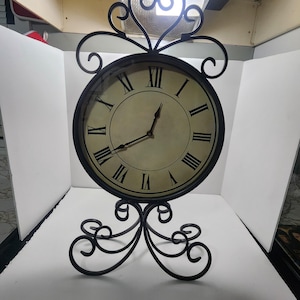 May include: A black metal table clock with a circular face and a cream-colored dial featuring black Roman numerals. The clock is supported by a decorative scrolled metal base and topped with a matching scrolled metal ornament, with two thin hands.