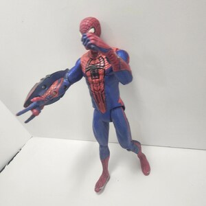 May include: A Spider-Man action figure in a dynamic pose. The figure is mainly blue and red, with a detailed spider-web pattern on the torso and mask. The figure has a web-shooter on the left arm.