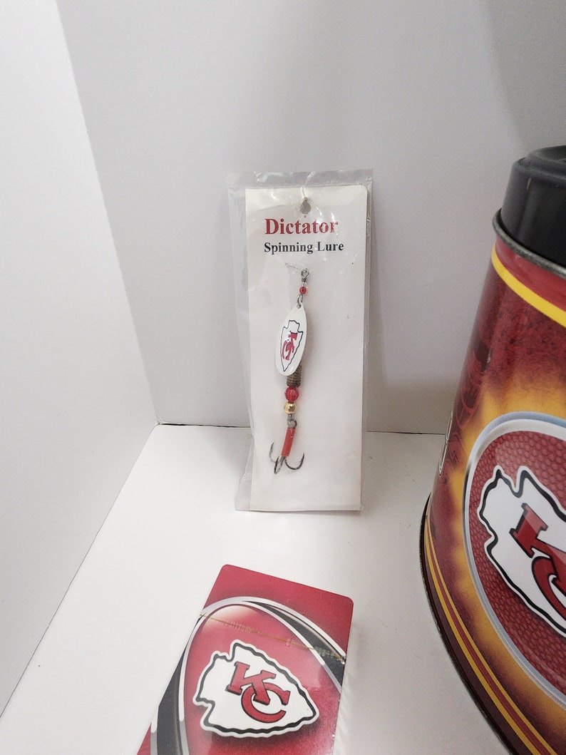 May include: A fishing lure in a clear plastic package with the text "Dictator Spinning Lure". The lure has a white oval body with a red and white logo. The lure has a red hook and red beads.