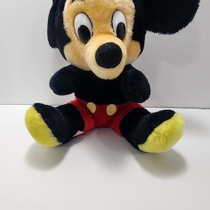 May include: A plush Mickey Mouse toy with a black body, ears, and arms. The face is tan with large white eyes and a black nose. The shorts are red with yellow buttons, and the feet are black with yellow soles. A classic Disney collectible.