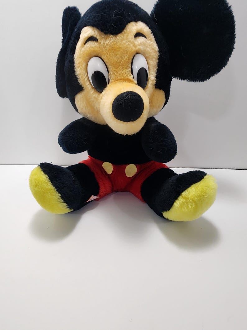 May include: A plush Mickey Mouse toy with a black body, ears, and arms. The face is tan with large white eyes and a black nose. The shorts are red with yellow buttons, and the feet are black with yellow soles. A classic Disney collectible.