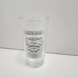 Könnte beinhalten: Ein klares Glas mit schwarzem und goldenem Text, darunter "WHITE SAPPHIRE" und "DISTILLED LONDON DRY GIN". Das Glas hat eine zylindrische Form und ist zum Servieren von Getränken geeignet. Der Text enthält auch "ESTD. 1770".
