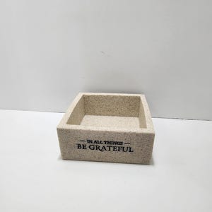 May include: A beige, rectangular decorative box with a textured surface. The box has the phrase "IN ALL THINGS - BE GRATEFUL" printed in black on the front. The box is empty and sits on a white surface.