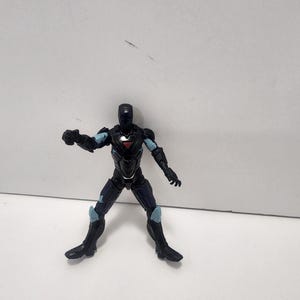 May include: Black and blue action figure of a superhero wearing a helmet and armor. The figure is posed in a fighting stance with one arm raised.