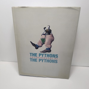May include: A hardcover book titled "The Pythons Autobiography by The Pythons" with a whimsical illustration. The cover features a figure in a shoe-shaped container, holding a bird. The book has a light gray background.