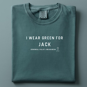 Op de afbeelding: Een opgevouwen tealkleurig T-shirt met de witte tekst "I WEAR GREEN FOR JACK", "CEREBRAL PALSY AWARENESS" en een witte lintafbeelding. Het shirt heeft een ronde hals en een label in het midden bovenaan.