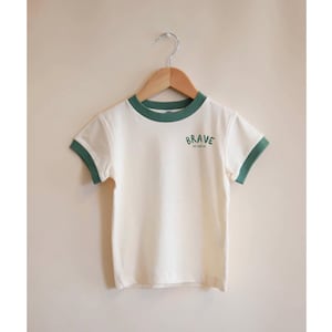 May include: Cream-colored t-shirt with green trim around the collar and sleeves. The word "BRAVE" is printed on the front in green, with the phrase "AS CAN BE" below it. The shirt is hanging on a wooden hanger.