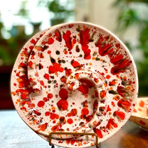 May include: White ceramic ashtray with a red and orange splatter pattern. The ashtray has three cigarette rests and a metal stand.