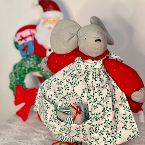 May include: A gray fabric mouse wearing a red and white dress with a green holly pattern. The mouse is sitting on a pair of red and white striped ice skates. The mouse is holding a small gray fabric mouse in its pocket.