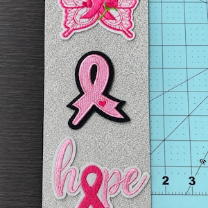 May include: Three embroidered patches on a silver glitter backing. The top patch is a pink butterfly with a ribbon body. The middle is a pink awareness ribbon with a black border and small heart. The bottom patch features the word hope in pink.