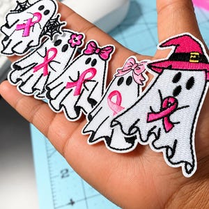 Puede incluir: Cinco parches bordados con forma de fantasma blanco que incluyen lazos rosas de concienciación sobre el cáncer de mama. Los fantasmas tienen bordes negros y accesorios como telarañas, una flor rosa, un lazo rosa y un sombrero de bruja rosa.