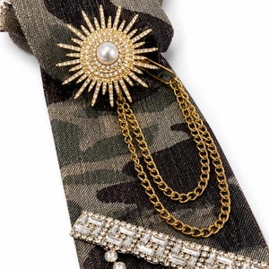 May include: A camouflage print necktie featuring a gold-tone sunburst brooch with a central faux pearl and clear rhinestones. Two gold-tone chains drape from the brooch. A rectangular bar with clear baguette crystals and a dangling rhinestone strand.