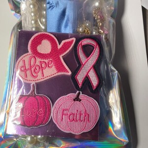 May include: A set of four embroidered patches in a holographic plastic bag. The patches include a pink ribbon with the word Hope, a pink awareness ribbon, and two pink pumpkins with the words Cure and Faith embroidered on them.