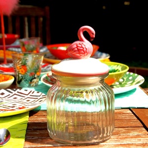 Könnte beinhalten: Ein klares Glasgefäß mit einem weißen Deckel und einer rosafarbenen Flamingofigur darauf. Das Gefäß steht auf einem Holztisch mit bunten Tellern und Schalen drumherum.