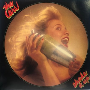 May include: A woman with blonde hair holds a silver cocktail shaker to her ear, her mouth open in a shout. The background is a bright orange color. The text "The Cars" is in the upper left corner and "Shake It Up" is in the lower right corner.