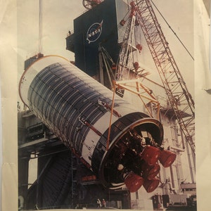 Op de afbeelding: Een zwart-witfoto van een tweede trap van een Saturn V-raket die wordt gehesen voor een testlancering in de Mississippi Test Facility van NASA. De foto is afkomstig van de Space Division van North American Rockwell Corporation.