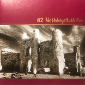 May include: A black and white photo of a crumbling stone building covered in vines. The building has a large archway and several windows. Two people are standing in front of the building. The text "U2 The Unforgettable Fire" is written in gold lettering on a red background.