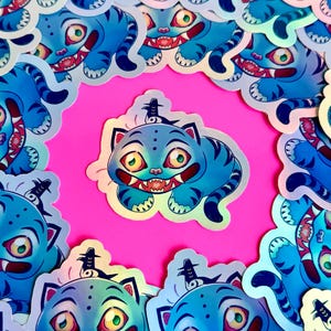 Peut inclure: Une collection d'autocollants représentant un chat de dessin animé avec une fourrure bleue, des rayures de tigre et un large sourire. Le chat a de grands yeux jaunes et est entouré d'une bordure holographique. Les autocollants sont posés sur un fond rose vif.