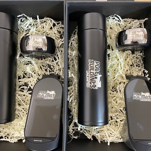 May include: Three black gift boxes with black and silver items inside. The boxes are filled with shredded paper. The items include a black water bottle, a black phone case, and a black ring. All of the items have the logo "POLE BUILDINGS UNLIMITED" printed on them.