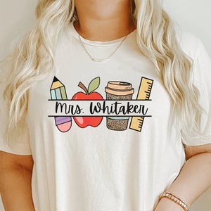 May include: A white t-shirt with a graphic design of a pencil, apple, coffee cup, and ruler. The text "Mrs. Whitaker" is printed on the shirt.