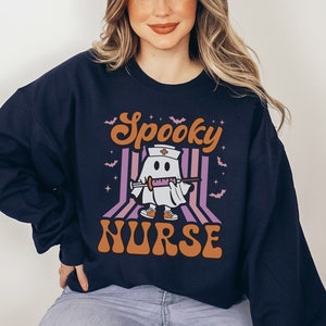 K&ouml;nnte beinhalten: Ein marineblauer Sweatshirt mit einem Aufdruck eines Geistes, der eine Krankenschwesterm&uuml;tze tr&auml;gt und eine Spritze h&auml;lt. Der Text "Spooky Nurse" ist in orangefarbenen und violetten Buchstaben gedruckt.