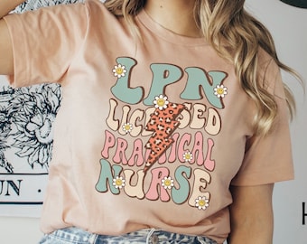 May include: A peach colored t-shirt with the text "LPN Licensed Practical Nurse" in a retro style font with a lightning bolt and flowers. The text is in shades of green, pink, and yellow.