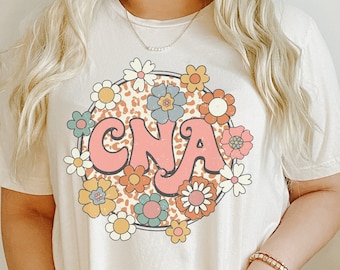 May include: A white t-shirt with a retro-style graphic of a floral wreath with the letters "CNA" in pink inside. The wreath is on a leopard print background.