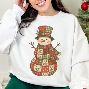 May include: White sweatshirt featuring a whimsical snowman design. The snowman has a patchwork body, a top hat, and a scarf in shades of red, green, and brown. The word "WHITE" is printed in white at the bottom.