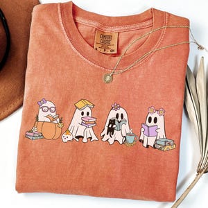 May include: A coral Comfort Colors sweatshirt featuring a graphic of four cartoon ghosts reading books. The ghosts are adorned with accessories like glasses and flowers. A gold necklace and a brown hat are also visible.