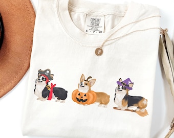 May include: Ivory-colored t-shirt featuring three corgis dressed in Halloween costumes. One corgi is a pirate, another is in a pumpkin costume, and the third is a witch. A gold necklace and a brown hat are also visible.