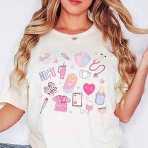 Puede incluir: Una camiseta blanca con un dise&ntilde;o rosa y azul que presenta una variedad de iconos m&eacute;dicos y relacionados con beb&eacute;s, incluyendo un estetoscopio, un biber&oacute;n, un chupete, una jeringa, un monitor de frecuencia card&iacute;aca, un pa&ntilde;al y la palabra "NICU".