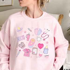 May include: Pink sweatshirt with a design of various medical symbols and the word "NICU" in white. The symbols include a stethoscope, a baby bottle, a pacifier, a syringe, a heart monitor, a thermometer, a baby footprint, a diaper, a baby in a crib, and a nurse's uniform.