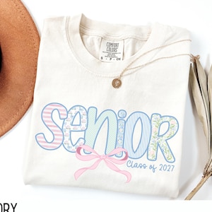 May include: Ivory-colored Comfort Colors t-shirt with the word "Senior" in pastel blue and pink patterns, a pink bow, and "Class of 2027" text. A gold necklace with a pendant and a brown hat are also visible.
