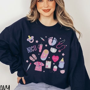 May include: A navy blue sweatshirt with a pink and blue graphic design featuring a variety of baby-related icons, including a pacifier, a syringe, a stethoscope, a baby bottle, and the word "NICU".