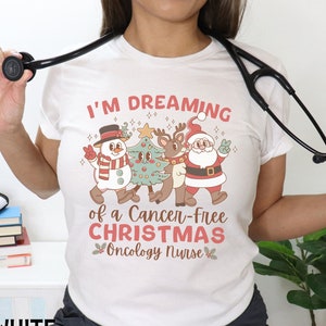May include: White t-shirt with a Christmas-themed design featuring a snowman, reindeer, and Santa Claus. The text reads "I'm Dreaming of a Cancer-Free Christmas Oncology Nurse".