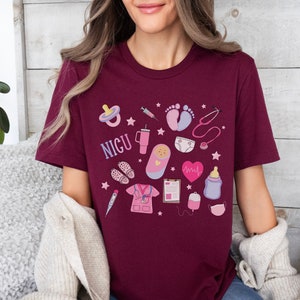 Puede incluir: Una camiseta color burdeos con un dise&ntilde;o gr&aacute;fico de varios s&iacute;mbolos m&eacute;dicos y la palabra "NICU". Los s&iacute;mbolos incluyen un estetoscopio, un biber&oacute;n, un chupete, una jeringa, un term&oacute;metro, un monitor de frecuencia card&iacute;aca, una huella de pie de beb&eacute;, un pa&ntilde;al y un beb&eacute; en un arrullo.