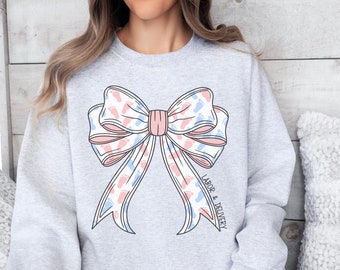 Könnte beinhalten: Ein hellgraues Sweatshirt mit einem großen Schleifendesign. Die Schleife ist mit rosa und blauen Babyfüßen verziert. Die Worte "LABOR & DELIVERY" sind vertikal auf der rechten Seite der Schleife aufgedruckt. Das Sweatshirt ist ein bequemer, lässiger Stil.