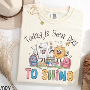 May include: Ivory-colored t-shirt with the text "Today Is Your Day TO SHINE" and a graphic of a smiling sun, a paper with an A+ grade, and two backpacks. A gold necklace and a hat are also visible.