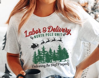 May include: A white t-shirt with a Christmas-themed design. The text on the shirt reads "Labor & Delivery North Pole Unit Delivering the Best Presents". The design also includes a cartoon image of Santa Claus in a sleigh with reindeer flying over a forest.