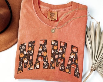 May include: A burnt orange t-shirt with the word "MAMA" printed in black with a fall-themed pumpkin and flower pattern inside the letters.