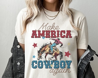 Peut inclure: T-shirt crème avec un motif d'un cow-boy sur un cheval sauvage, et le texte "Make AMERICA COWBOY again" en rouge et bleu. Le design comprend des étoiles et des lettres en forme de corde.