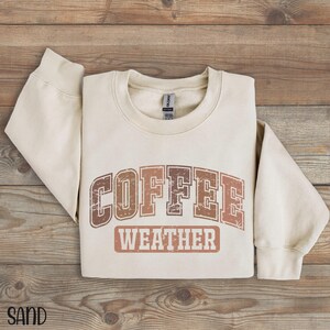May include: Sand-colored sweatshirt with the words "COFFEE WEATHER" in a distressed, brown font. The sweatshirt is folded on a wooden surface. The word "SAND" is printed in the bottom left corner.