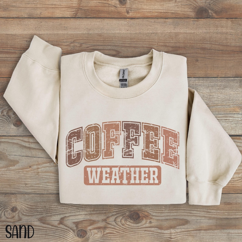 May include: Sand-colored sweatshirt with the words "COFFEE WEATHER" in a distressed, brown font. The sweatshirt is folded on a wooden surface. The word "SAND" is printed in the bottom left corner.