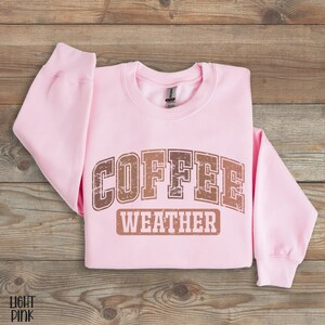May include: A light pink sweatshirt with the words "COFFEE WEATHER" printed in brown, distressed lettering. The sweatshirt is folded on a wooden surface. The text is in a vintage, collegiate style.