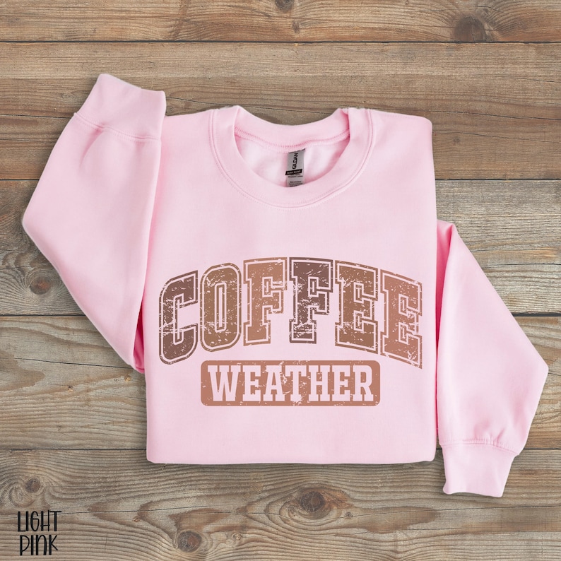 May include: A light pink sweatshirt with the words "COFFEE WEATHER" printed in brown, distressed lettering. The sweatshirt is folded on a wooden surface. The text is in a vintage, collegiate style.