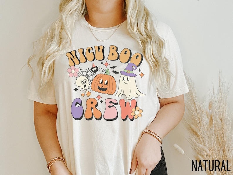 May include: A white t-shirt with a retro-style graphic that says "NICU BOO CREW" with a ghost, pumpkin, and spider. The graphic is in shades of orange, purple, pink, and yellow.