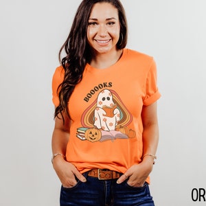 May include: Orange t-shirt featuring a ghost reading a book with the text "BOO-OOOKS" in a rainbow arch. The ghost is wearing a floral pattern and there are pumpkins and books on the ground.